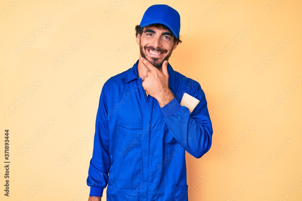 Handsome young man with curly hair and bear wearing builder jumpsuit uniform looking confident at the camera smiling with crossed arms and hand raised on chin. thinking positive.