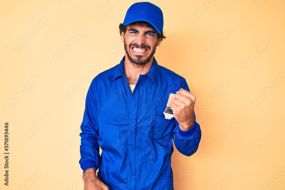 Handsome young man with curly hair and bear wearing builder jumpsuit uniform angry and mad raising fist frustrated and furious while shouting with anger. rage and aggressive concept.