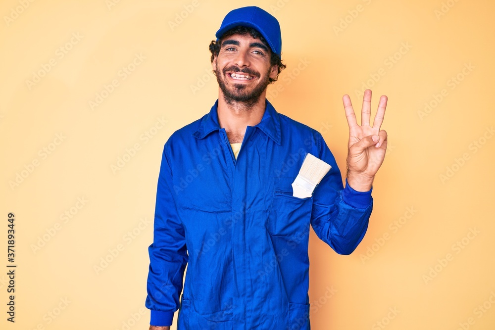 Handsome young man with curly hair and bear wearing builder jumpsuit uniform showing and pointing up with fingers number three while smiling confident and happy.