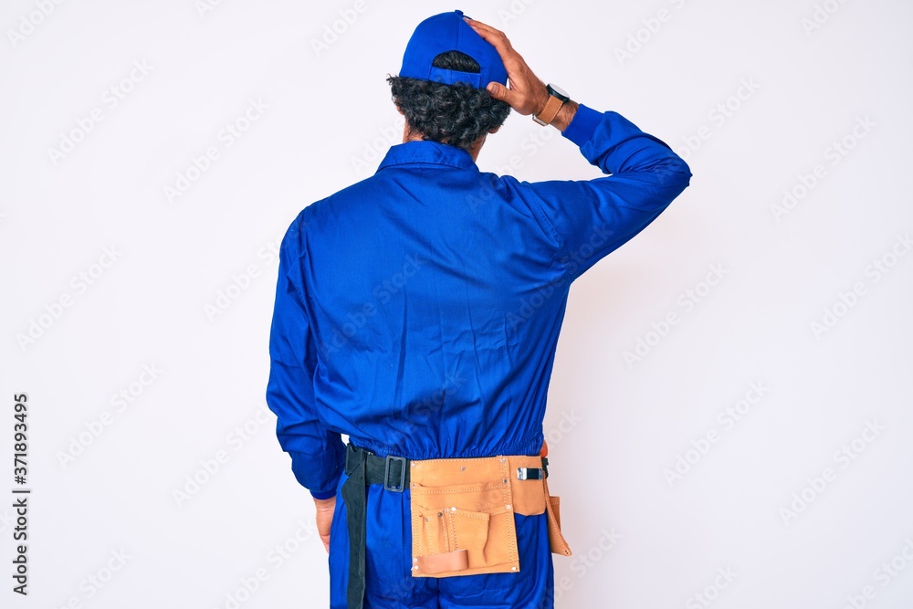 Handsome young man with curly hair and bear weaing handyman uniform backwards thinking about doubt with hand on head Handsome young man with curly hair and bear weaing handyman uniform backwards thinking about doubt with hand on head