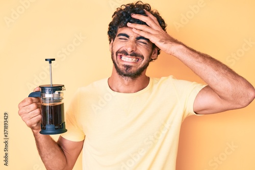 Handsome young man with curly hair and bear holding french coffee maker stressed and frustrated with hand on head, surprised and angry face