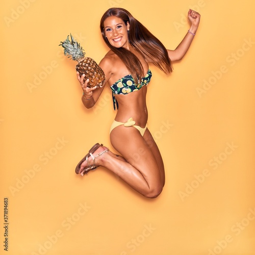 Young beautiful hispanic woman wearing bikini smiling happy. Jumping with smile on face holding pineapple over isolated yellow background.