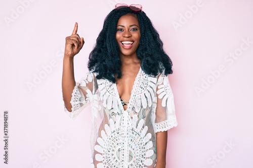 Beautiful african woman wearing summer dress and sunglasses pointing finger up with successful idea. exited and happy. number one.