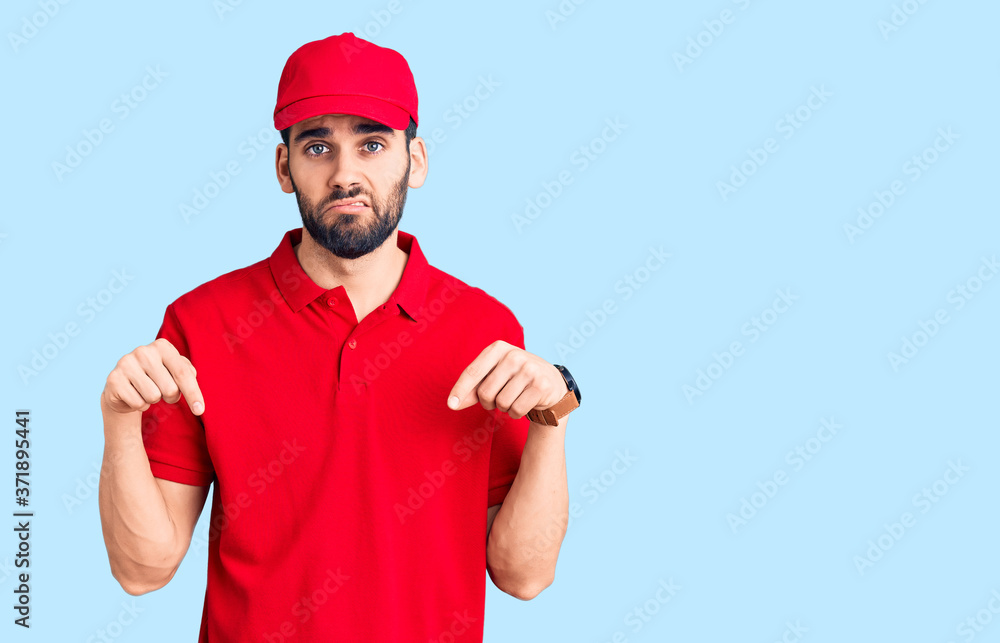 Young handsome man with beard wearing delivery uniform pointing down looking sad and upset, indicating direction with fingers, unhappy and depressed.