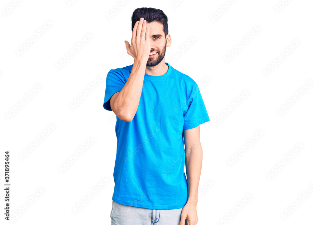 Young handsome man with beard wearing casual t-shirt covering one eye with hand, confident smile on face and surprise emotion. Young handsome man with beard wearing casual t-shirt covering one eye with hand, confident smile on face and surprise emotion.