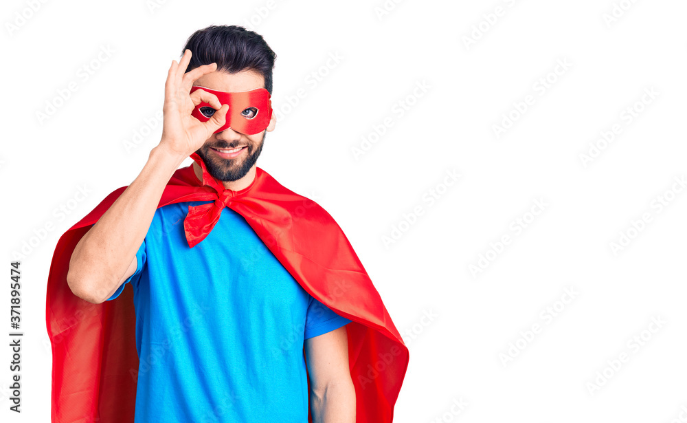 Young handsome man with beard wearing super hero costume doing ok gesture with hand smiling, eye looking through fingers with happy face. Young handsome man with beard wearing super hero costume doing ok gesture with hand smiling, eye looking through fingers with happy face.
