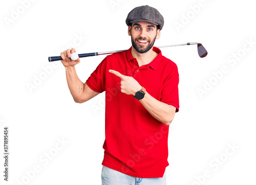 Young handsome man with beard playing golf holding club and ball smiling happy pointing with hand and finger