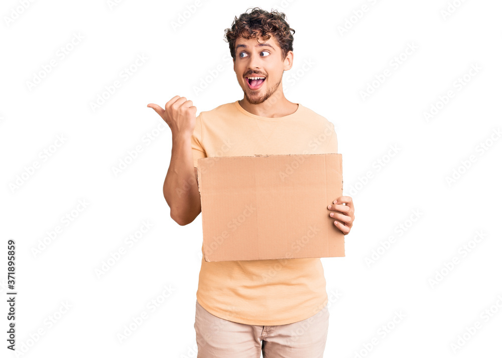 Young handsome man with curly hair holding blank cardboard empty banner pointing thumb up to the side smiling happy with open mouth Young handsome man with curly hair holding blank cardboard empty banner pointing thumb up to the side smiling happy with open mouth