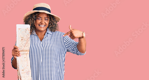 Young african american plus size woman wearing summer hat holding city map pointing finger to one self smiling happy and proud