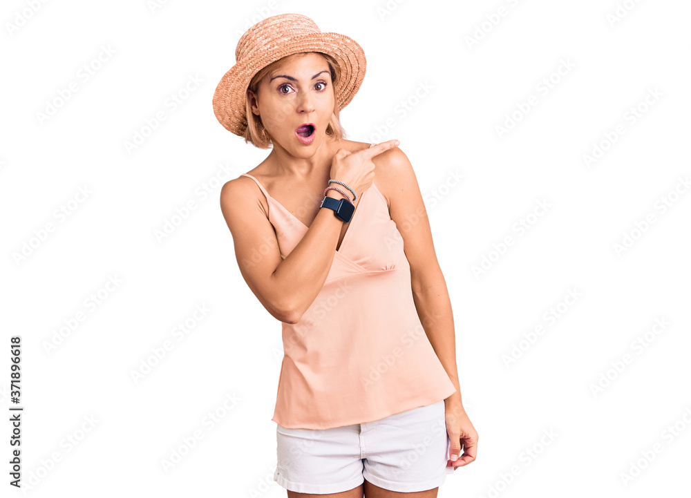 Young blonde woman wearing summer hat surprised pointing with finger to the side, open mouth amazed expression.