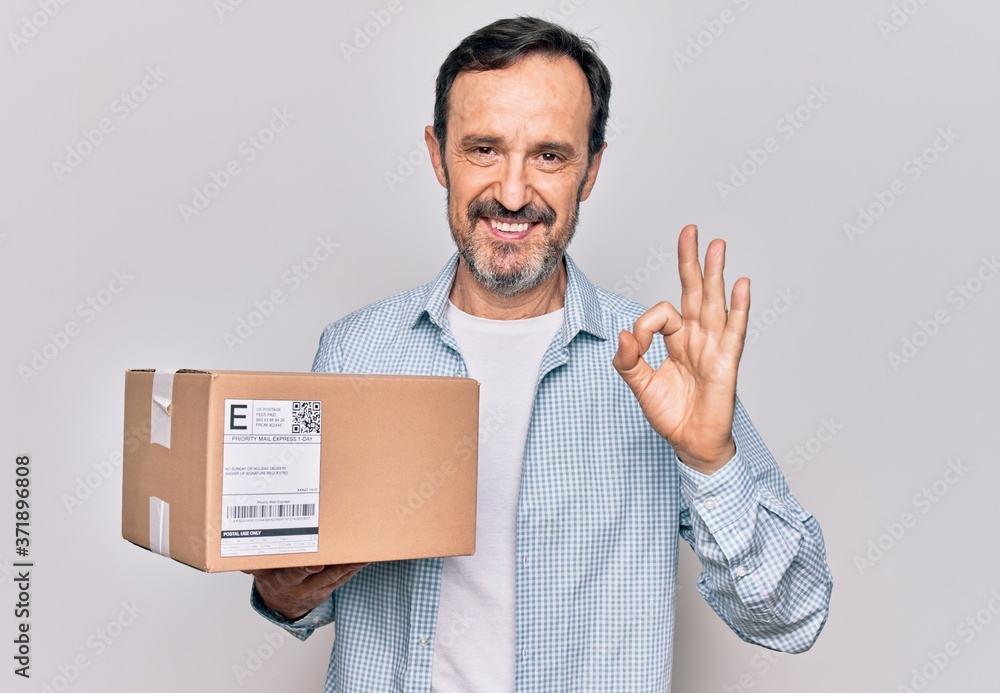 Middle age handsome man holding package over isolated white backgrounds doing ok sign with fingers, smiling friendly gesturing excellent symbol Middle age handsome man holding package over isolated white backgrounds doing ok sign with fingers, smiling friendly gesturing excellent symbol