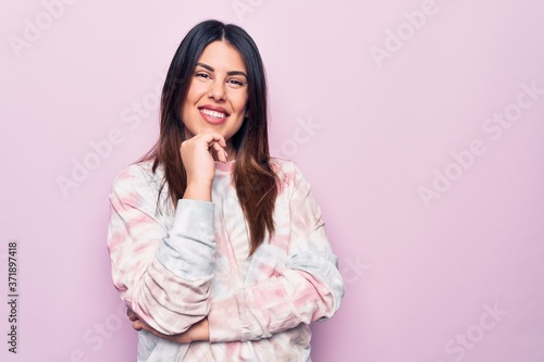 Young beautiful brunette woman wearing casual dyed sweatshirt over isolated pink background smiling looking confident at the camera with crossed arms and hand on chin. Thinking positive. © Krakenimages.com