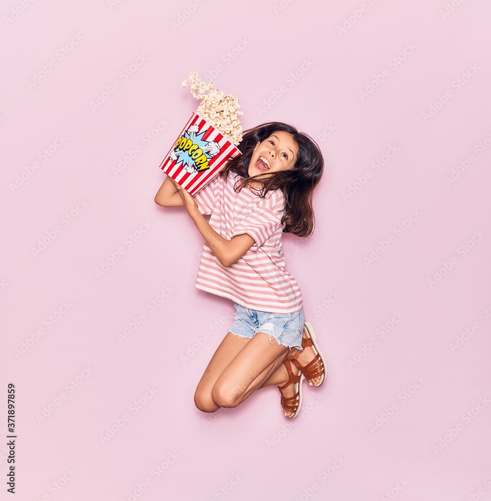 Adorable hispanic child girl surprised with open mouth holding bucket of popcorn jumping over isolated pink background Adorable hispanic child girl surprised with open mouth holding bucket of popcorn jumping over isolated pink background