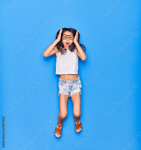 Adorable hispanic child girl wearing casual clothes and glasses surprised with open mouth. Jumping with hands on face  over isolated blue background