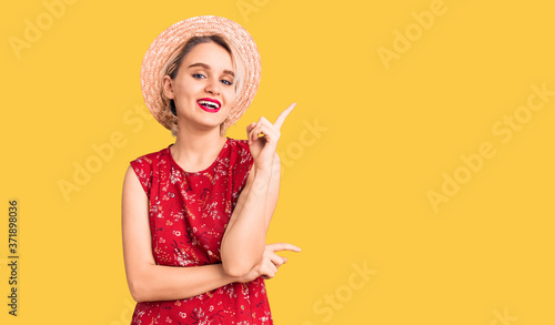 Young beautiful blonde woman wearing summer hat with a big smile on face, pointing with hand and finger to the side looking at the camera.