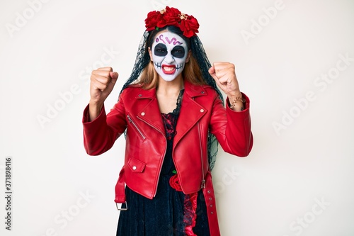 Woman wearing day of the dead costume over white angry and mad raising fists frustrated and furious while shouting with anger. rage and aggressive concept.