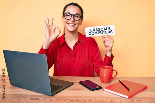 Young brunette woman using laptop holding exascale computers banner doing ok sign with fingers, smiling friendly gesturing excellent symbol photo