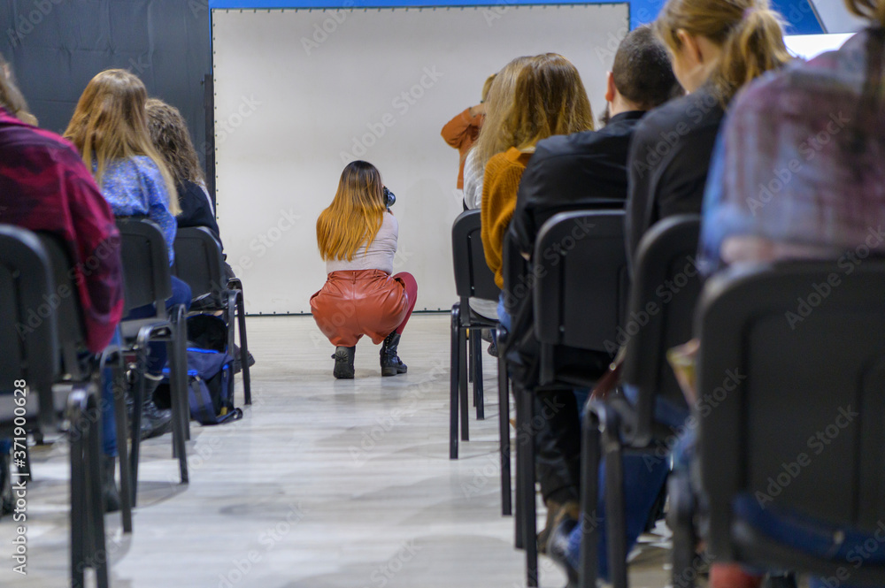Obraz premium The girl in the aisle between the chairs. Taking pictures with the camera. View from the back. Concept - seminar, workshop. Selective focus.