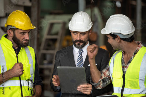 Factory manager in suit using laptop and talking with industrial engineer ,technician man. They wear helmet or hardhat with safety jackets inside industry manufacturing Facility/Discussion working