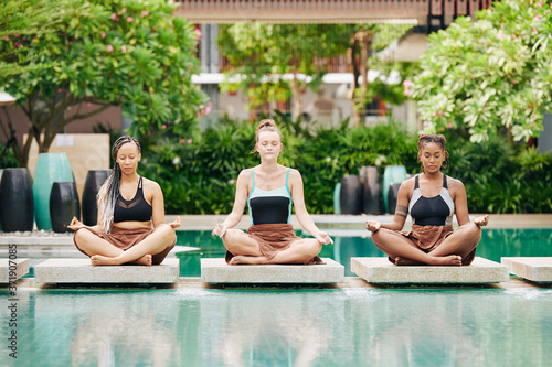 Pretty young women in swimsuits sitting in lotus position and doing breathing exercise to calm down and meditate