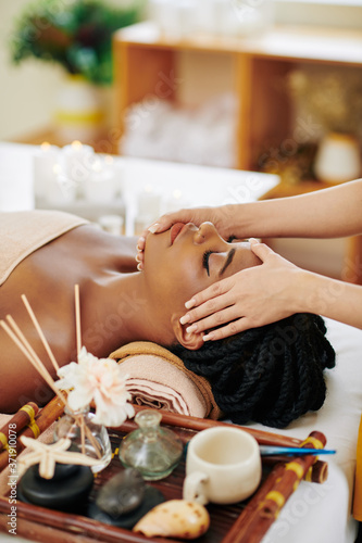 Beautiful young Black woman enjoying face and head massage in spa salon when lying on bed next to tray with aroma oils photo