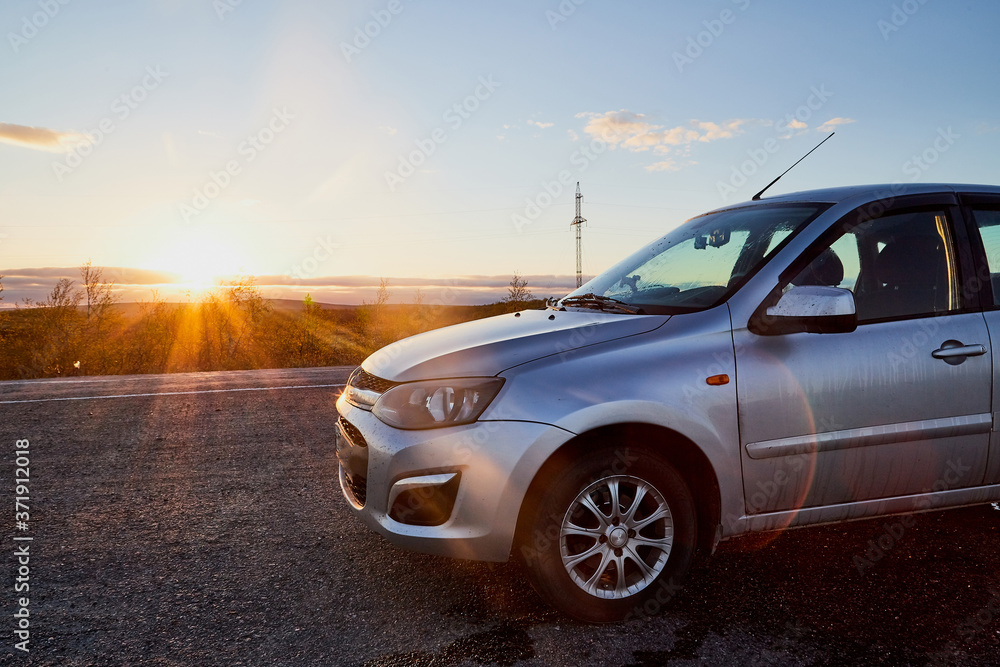 Small car standing on the side of asphalt road and sunset or sunrise with rays of sun behind it.