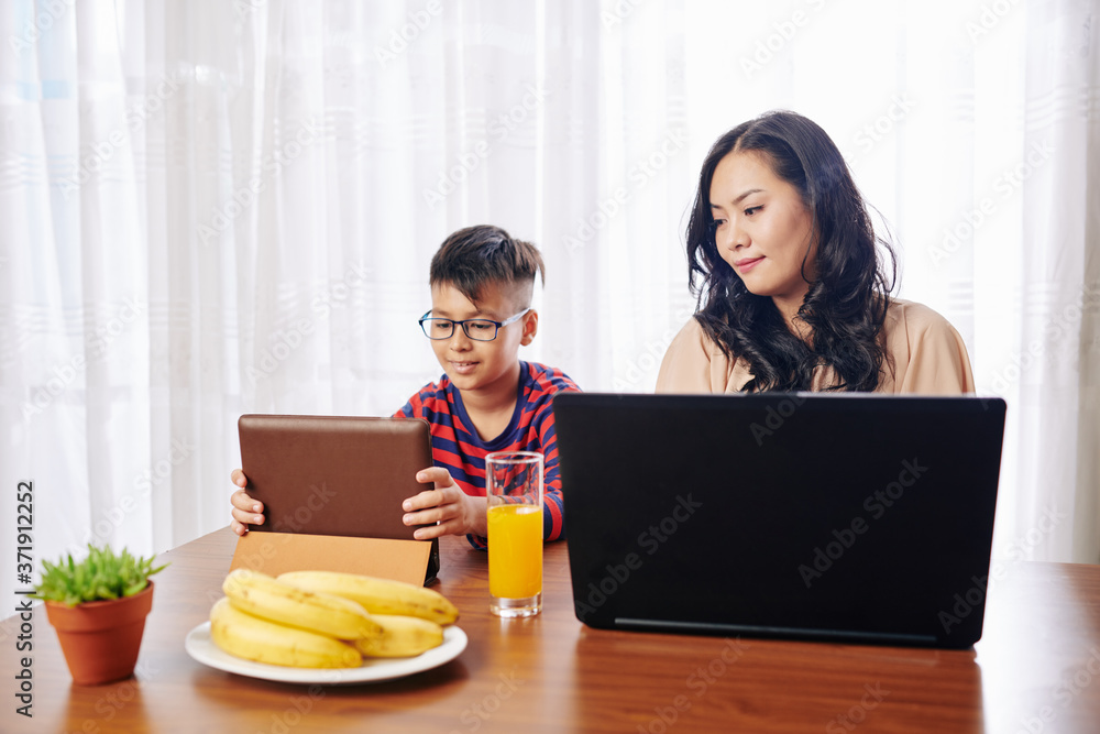 Beautiful young woman at kitchen table working on laptop, her son sitting nearby and watching educational video on digital tablet Beautiful young woman at kitchen table working on laptop, her son sitting nearby and watching educational video on digital tablet
