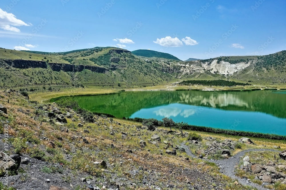 Beautiful mountain lake Nar. An emerald round-shaped reservoir in the crater of an extinct volcano. Summer sunny day, blue sky, light clouds. Reflection in water. Cappadocia. Turkey.
