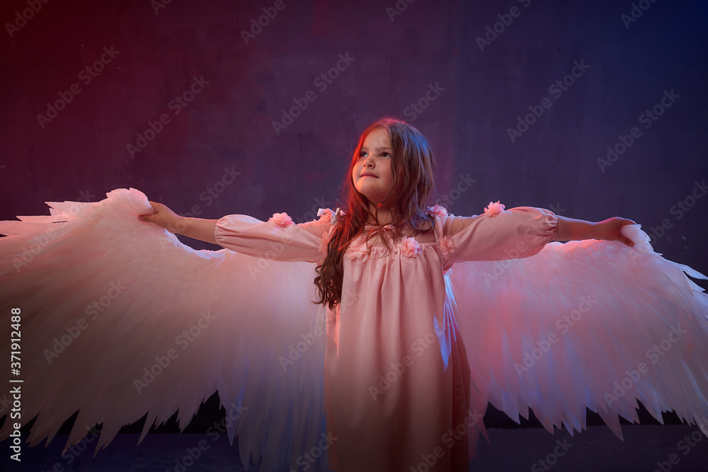 Naklejka premium Small young model girl with white wings posing and dancing in dark black studo during photoshoot with flour or dust and colour light. Actress in performance about struggle between good and evil.