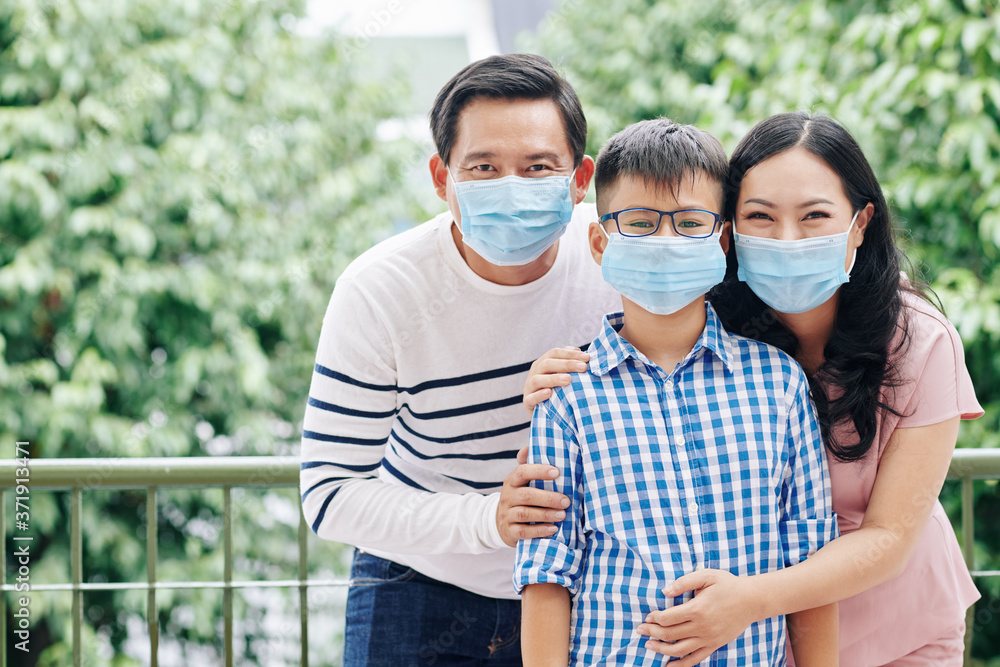 Cheerful Asian family of three in medical masks standing outdoors and looking at camera
