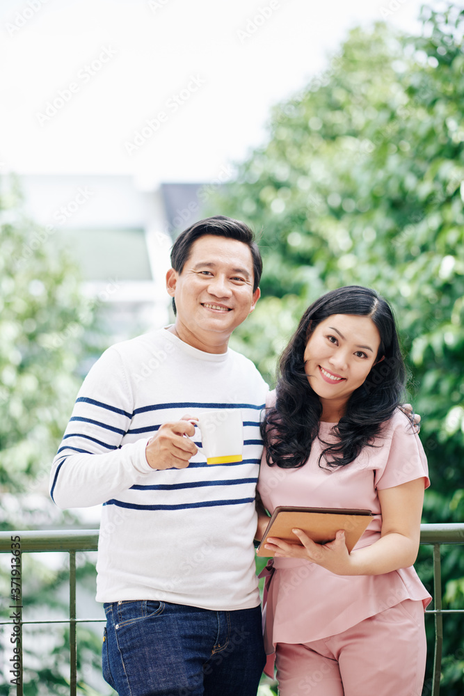 Portrait of cheerful Vietnamese husband and wife standing outdoors with morning coffee and digital tablet Portrait of cheerful Vietnamese husband and wife standing outdoors with morning coffee and digital tablet