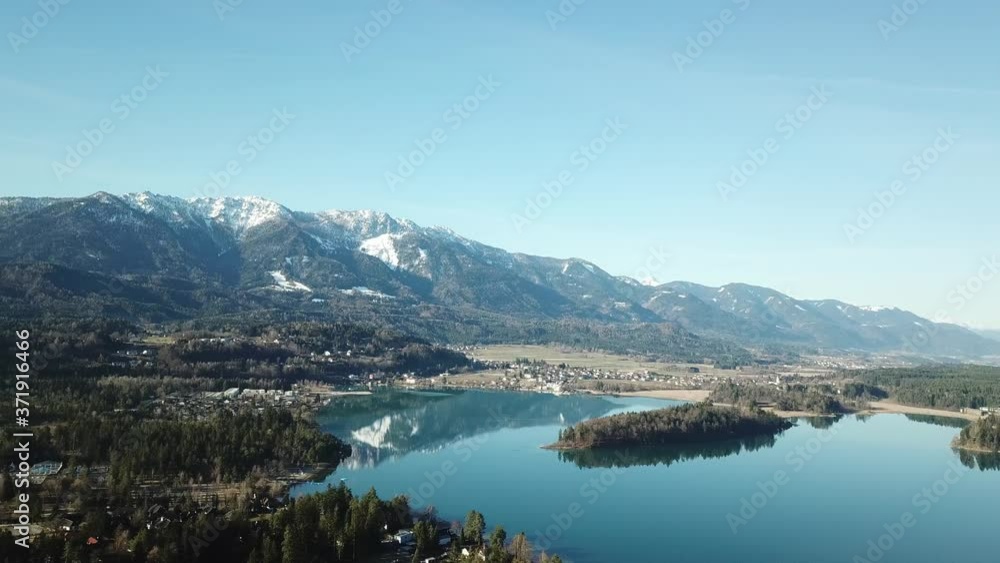 A drone flight above the Faaker See in Austria. There are high Alpine ranges around, reflecting in the crystal clear and calm surface of the lake. Small villages around the lake. Snow capped mountains A drone flight above the Faaker See in Austria. There are high Alpine ranges around, reflecting in the crystal clear and calm surface of the lake. Small villages around the lake. Snow capped mountains