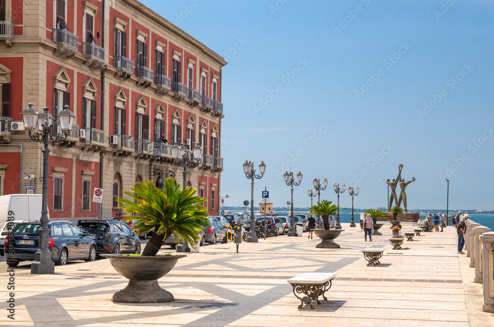 Naklejka premium Taranto, Italy - May 7, 2018: Embankment promenade with building, street lights, banches, trees and sailor monument in front of blue sky near sea channel in historical centre of city, Puglia (Apulia)