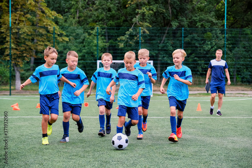 Fototapeta Naklejka Na Ścianę i Meble -  Kids running and kicking soccer ball on warming before football training session. Coach in the background watches the training process