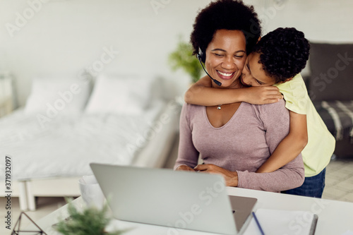 African American son embracing his mother who is working on a computer at home.