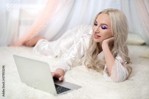 Glamorous blonde with a laptop. A beautiful girl in the bedroom with a laptop smiling communicates online .