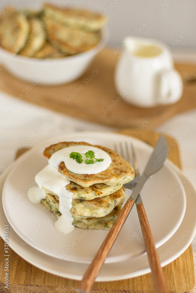 Zucchini pancakes with herbs on a light plate and a wooden board with cutlery, in the background a gravy boat with sour cream and a bowl of pancakes close-up Zucchini pancakes with herbs on a light plate and a wooden board with cutlery, in the background a gravy boat with sour cream and a bowl of pancakes close-up