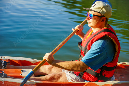 the guy is sailing on a red kayak, wearing a life jacket with glasses and a cap. Holds a paddle in his hand