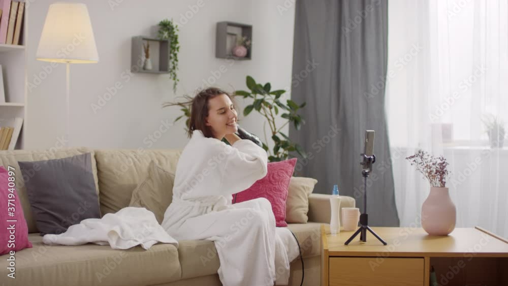 Beautiful Caucasian woman wearing white bath robe after taking morning shower drying her hair using hairdryer in front of smartphone camera Beautiful Caucasian woman wearing white bath robe after taking morning shower drying her hair using hairdryer in front of smartphone camera