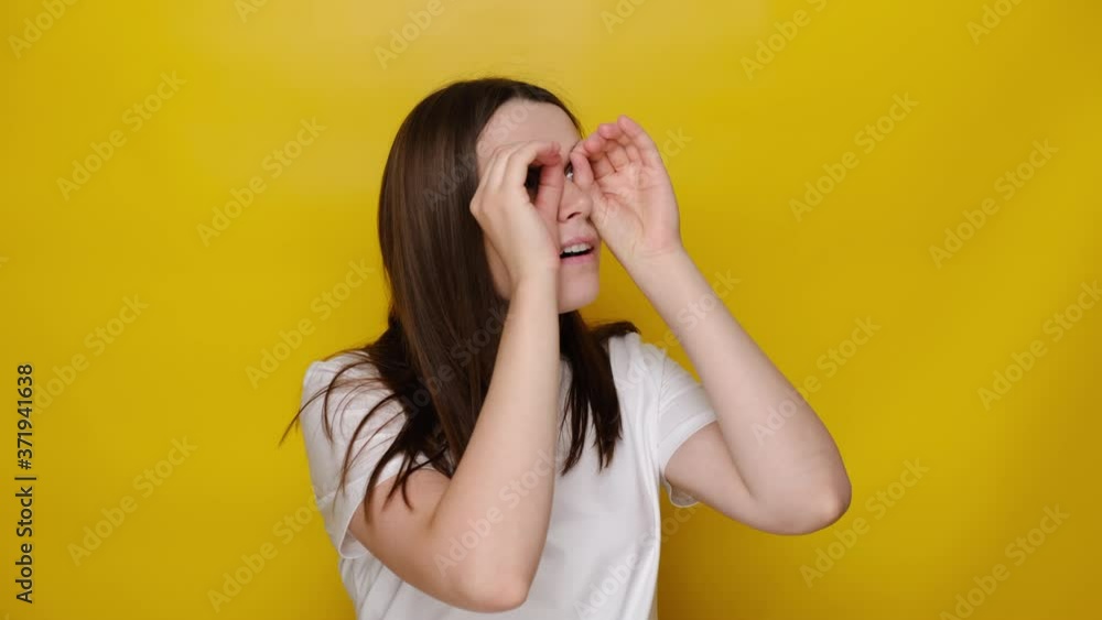 Curious young girl observing distant horizon, watching through binoculars hand gesture, brunette woman expressing amazement great surprise when looking at camera, isolated on yellow studio background Curious young girl observing distant horizon, watching through binoculars hand gesture, brunette woman expressing amazement great surprise when looking at camera, isolated on yellow studio background
