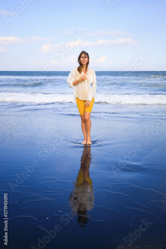 Beautiful young woman walking on black sand beach. Caucasian woman wearing yellow sportswear and white blouse. Romantic concept. Water reflection. Copy space.