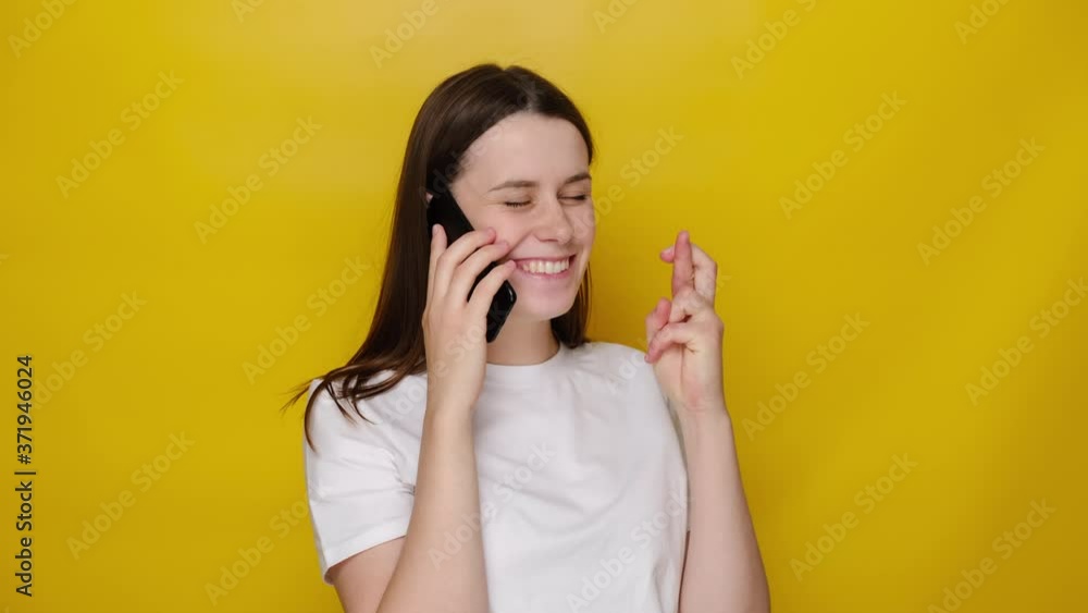 Cheerful young woman talking on smartphone, isolated on yellow studio background, gesturing finger crossed smiling with hope and eyes closed, dressed in white t-shirt. Luck and superstition concept Cheerful young woman talking on smartphone, isolated on yellow studio background, gesturing finger crossed smiling with hope and eyes closed, dressed in white t-shirt. Luck and superstition concept