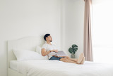 Asian man lying on bed work on his laptop in cozy white bedroom.