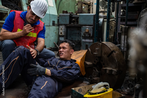 Young Asian man worker had a leg accident lying on the floor in metal lathe factory. Engineer supervisor talking on walkie talkie for help. Factory industrial safety and business insurance concept.