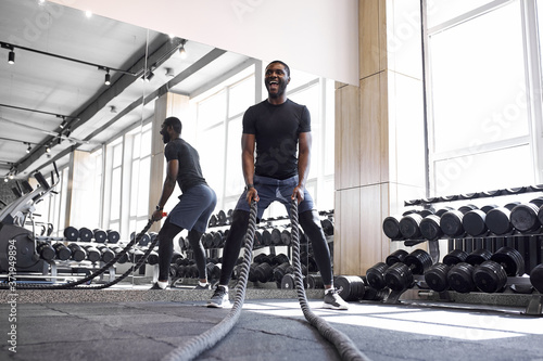 african-american man doing battle rope exercises in fitness gym, young muscular male performing crossfit training