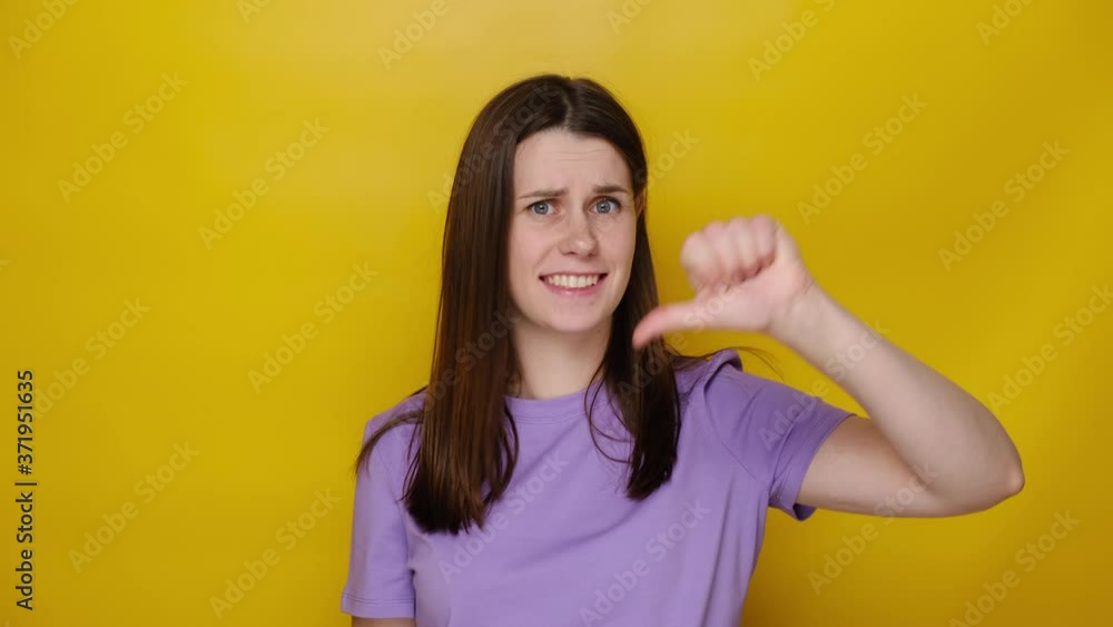 Dissatisfied young female showing thumbs down gesture, unhappy brunette woman customer giving negative feedback on product or service, wears purple t-shirt, isolated on yellow studio background Dissatisfied young female showing thumbs down gesture, unhappy brunette woman customer giving negative feedback on product or service, wears purple t-shirt, isolated on yellow studio background