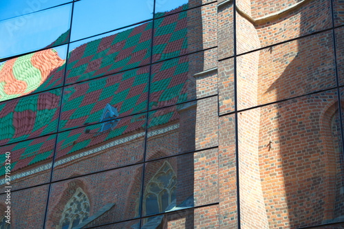 Reflection of an old, gothic church with a multi-colored roof and beautiful architectural forms in the glass of a new modern building against a blue sky, wallpaper for the screen