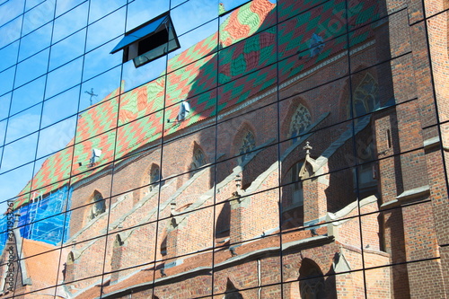 Reflection of an old, Gothic church with a multi-colored roof and beautiful architectural forms in the glass of a new modern building against a blue sky with an open window, wallpaper for the screen