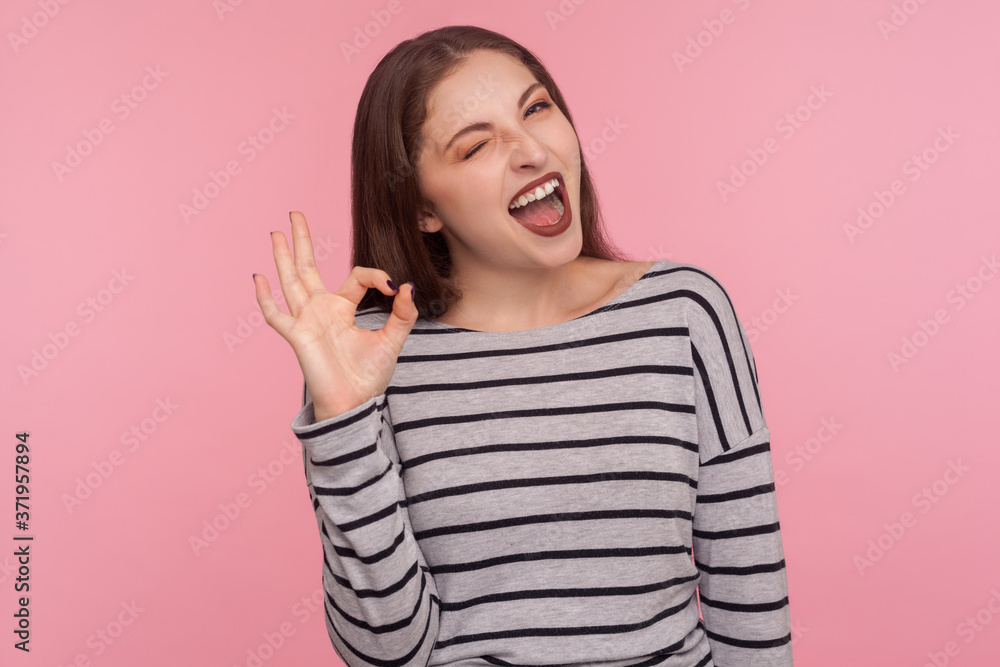 I am okay, approval. Portrait of satisfied happy young woman in striped sweatshirt doing ok gesture and winking to camera, agree with suggestion. indoor studio shot isolated on pink background I am okay, approval. Portrait of satisfied happy young woman in striped sweatshirt doing ok gesture and winking to camera, agree with suggestion. indoor studio shot isolated on pink background
