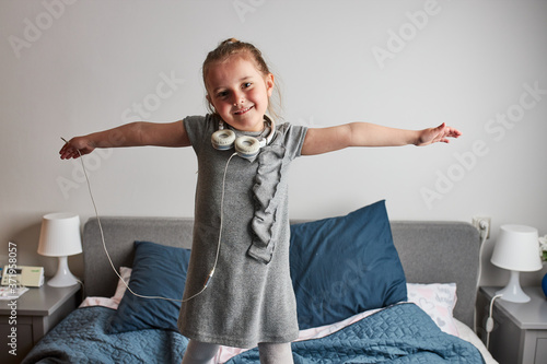 Little girl singing holding headphones cord imitating herself a real singer. Child having fun jumping dancing listening to music on bed in bedroom at home
